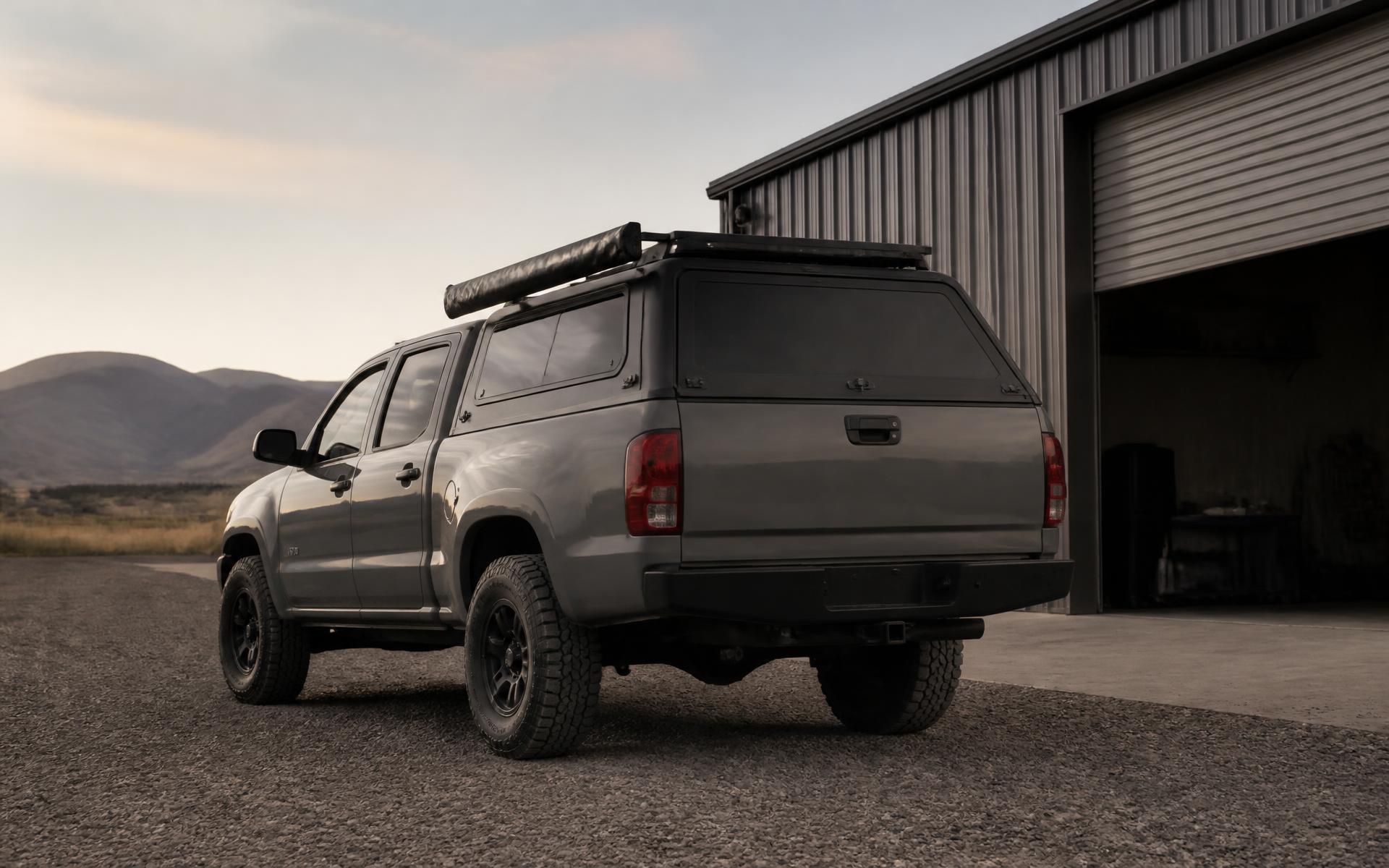 Rear three-quarter view of a completed overland pickup build parked on the gravel apron outside a workshop at golden hour, canopy fitted, low-profile roof rack with an awning, modest lift and all-terrain tires