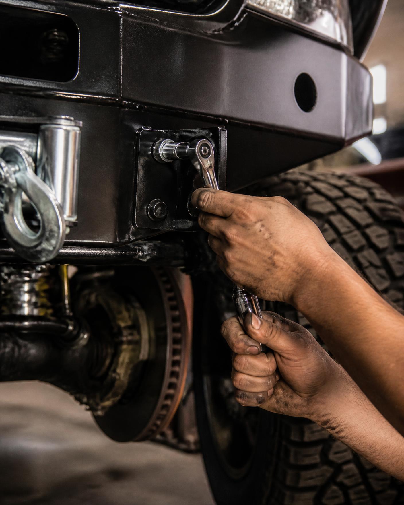 Close view of a mechanic's hands torquing a bolt on a newly fitted steel front bumper and winch mount, brake rotor and all-terrain tire visible behind, workshop light on bare metal and black powder coat