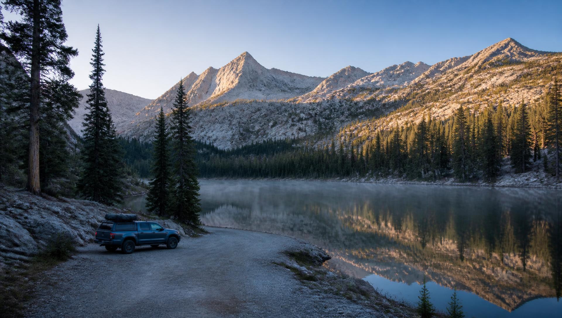 A modestly equipped overland pickup parked at a gravel pullout beside a still alpine lake at dawn, mist on the water, granite peaks and conifer forest filling the frame
