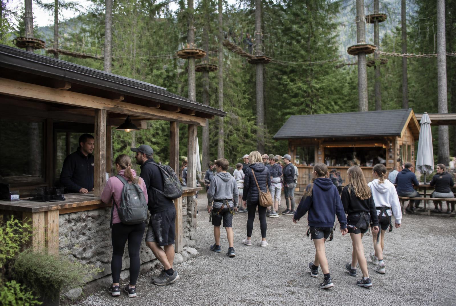 Busy adventure park base area with guests checking in at a timber cabin, a small food kiosk, picnic tables and aerial course platforms in the conifer canopy above