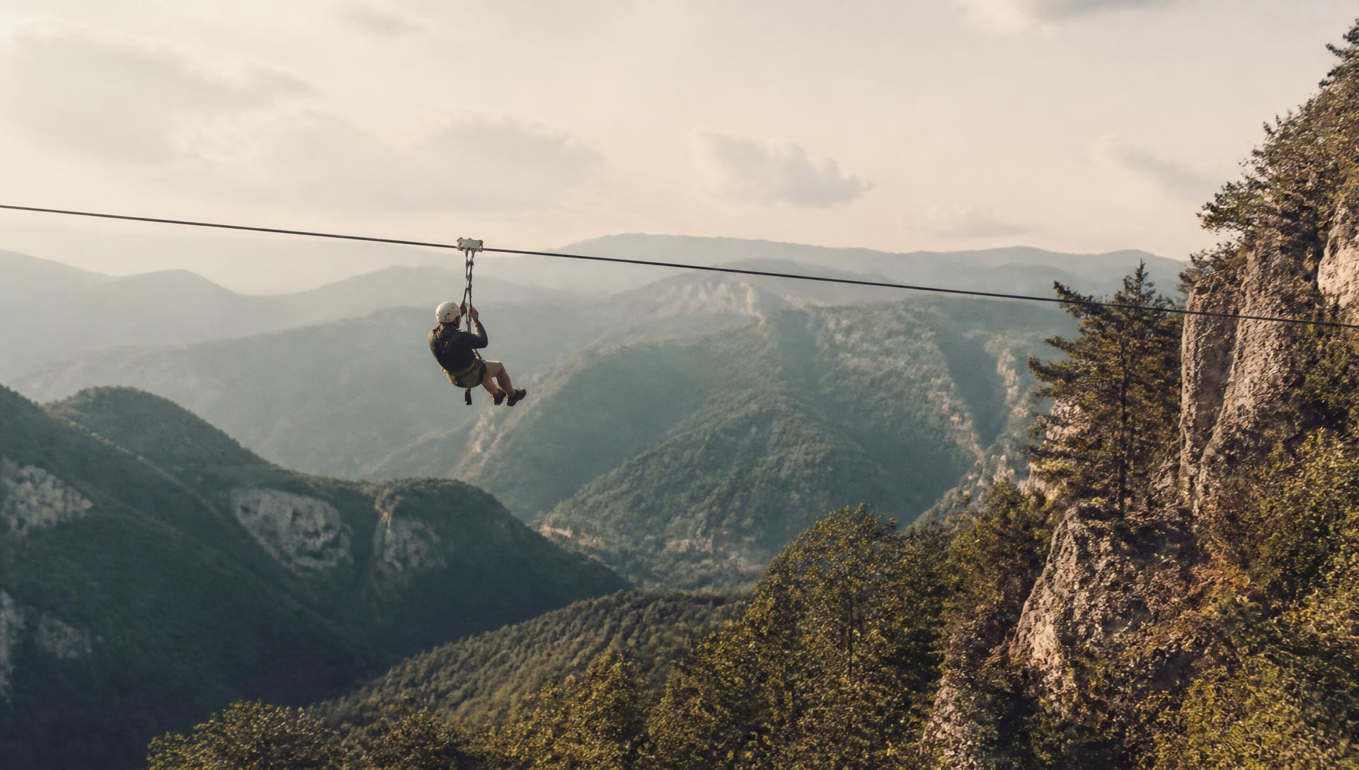 A single person in a helmet and harness riding a long zipline high above a forested valley, with layered ridgelines and afternoon haze behind them