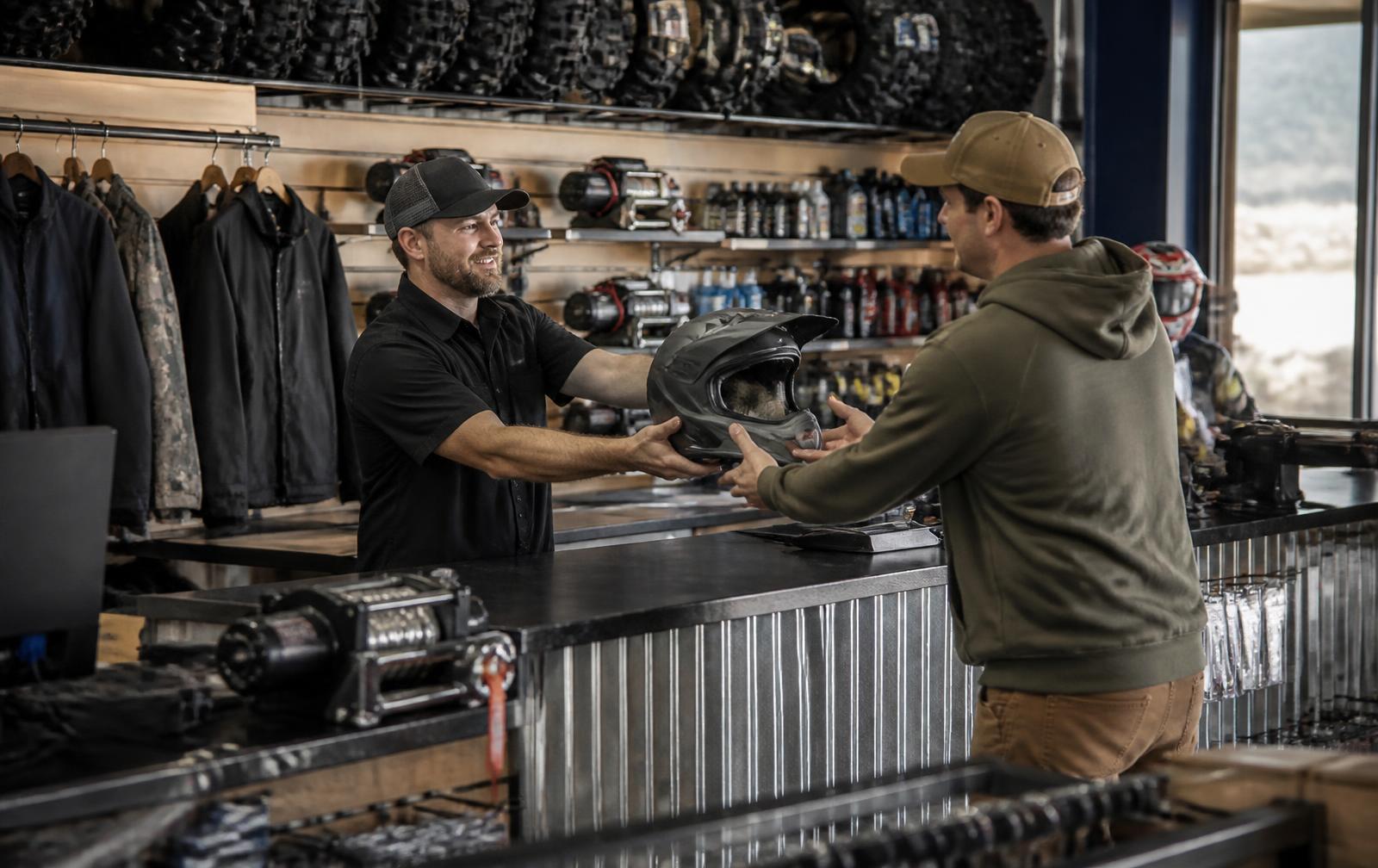 Parts counter employee handing a helmet to a customer, with riding gear, tires and winches on the shelves behind