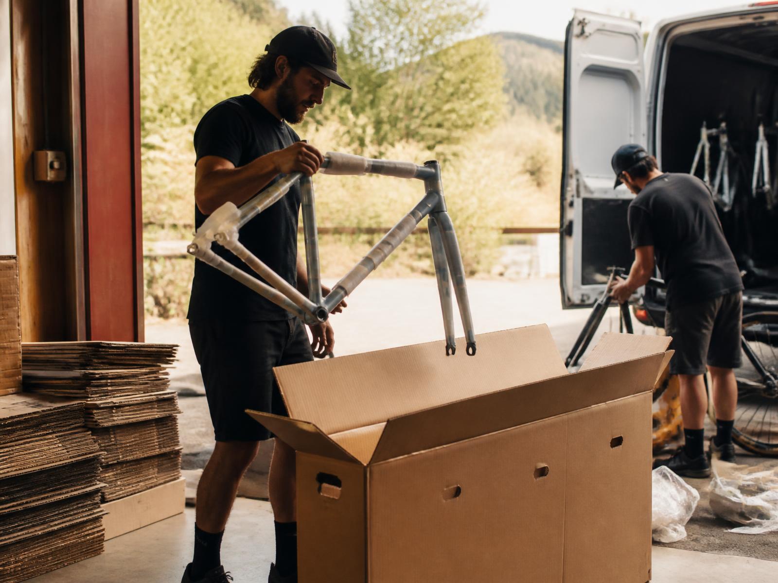 Two bike shop staff members unloading and assembling new bikes from cartons beside a delivery van at an open shop door