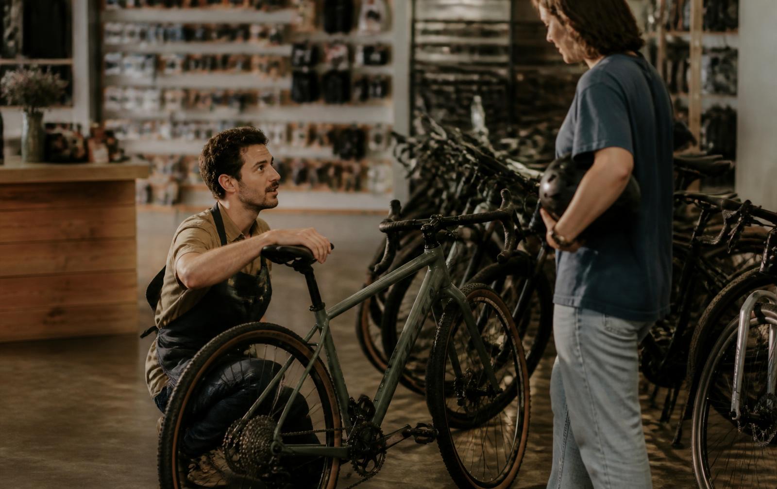 Bike shop staff member crouching beside a gravel bike on the sales floor, talking with a customer holding a helmet