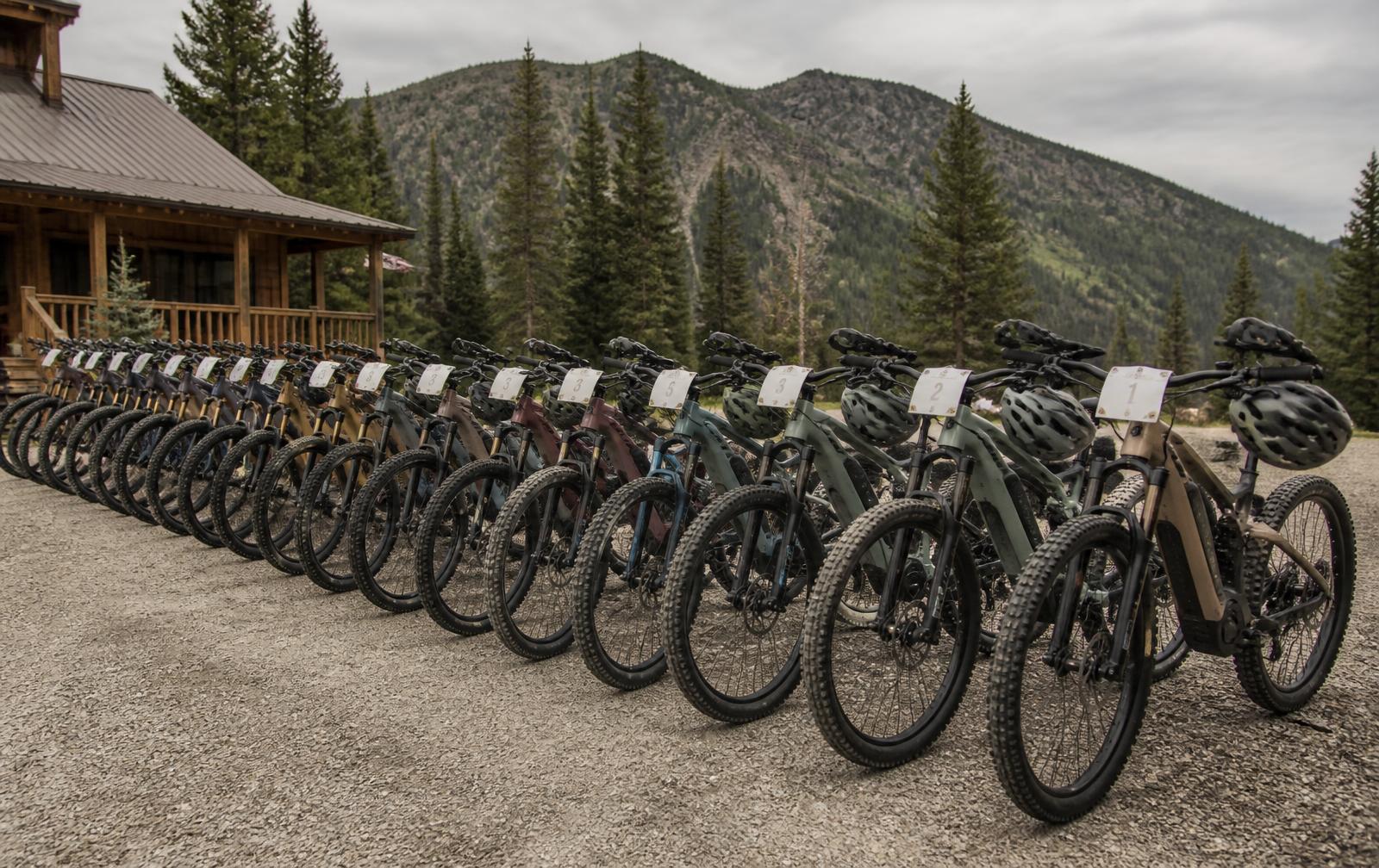 A long row of numbered rental mountain bikes and e-bikes staged on a gravel forecourt before a tour departure, helmets hung on some handlebars, pine forest and a mountain ridge behind under soft morning light