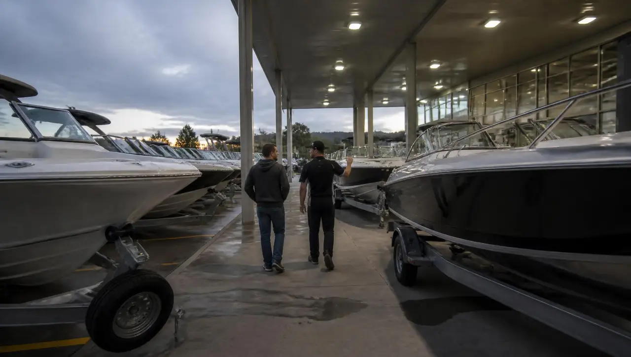 Salesperson walking a customer between rows of new boats on trailers outside a boat dealership showroom at dusk