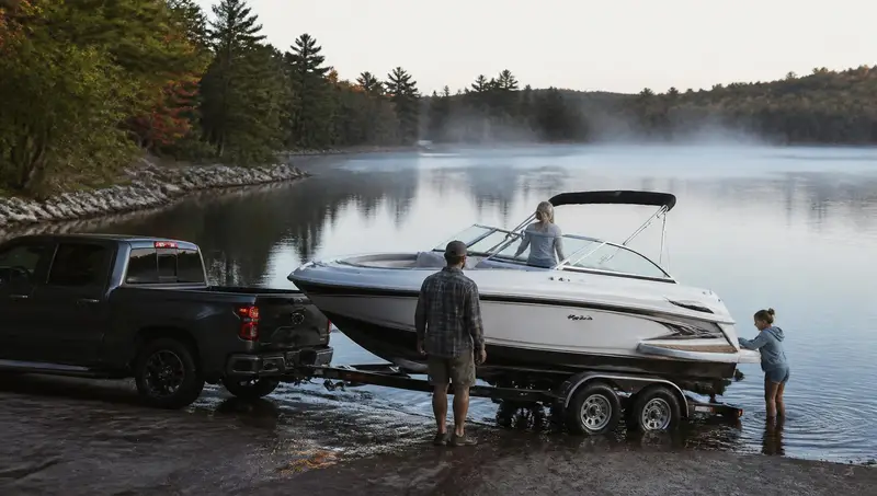 Family launching a newly delivered boat from a trailer at a quiet lake ramp in early morning light