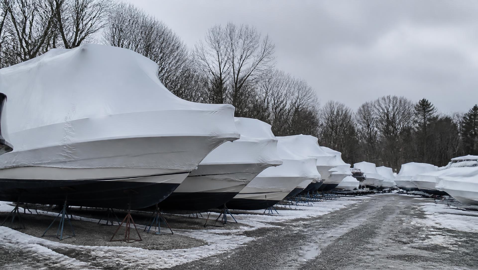 Rows of shrink-wrapped boats on stands in a winter storage yard behind a dealership under overcast sky