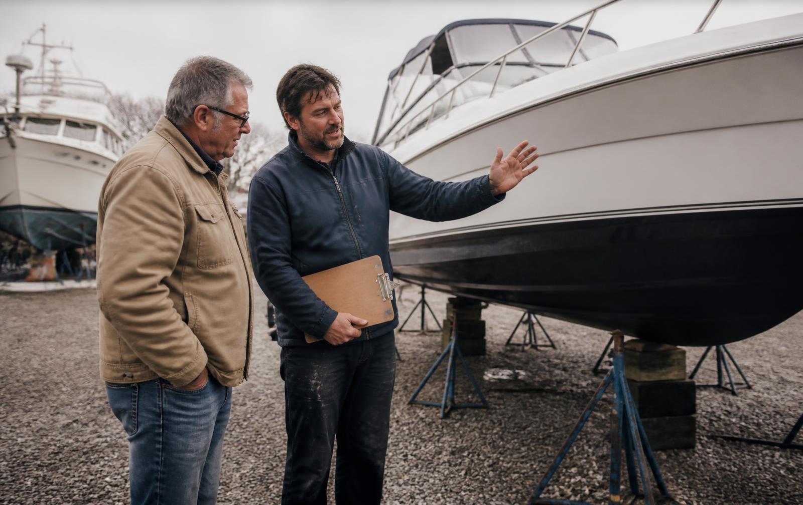 A boat owner in a canvas jacket and a technician holding a clipboard standing together beside a freshly serviced motorboat on stands in a gravel boatyard, the technician gesturing along the hull as he explains the completed work
