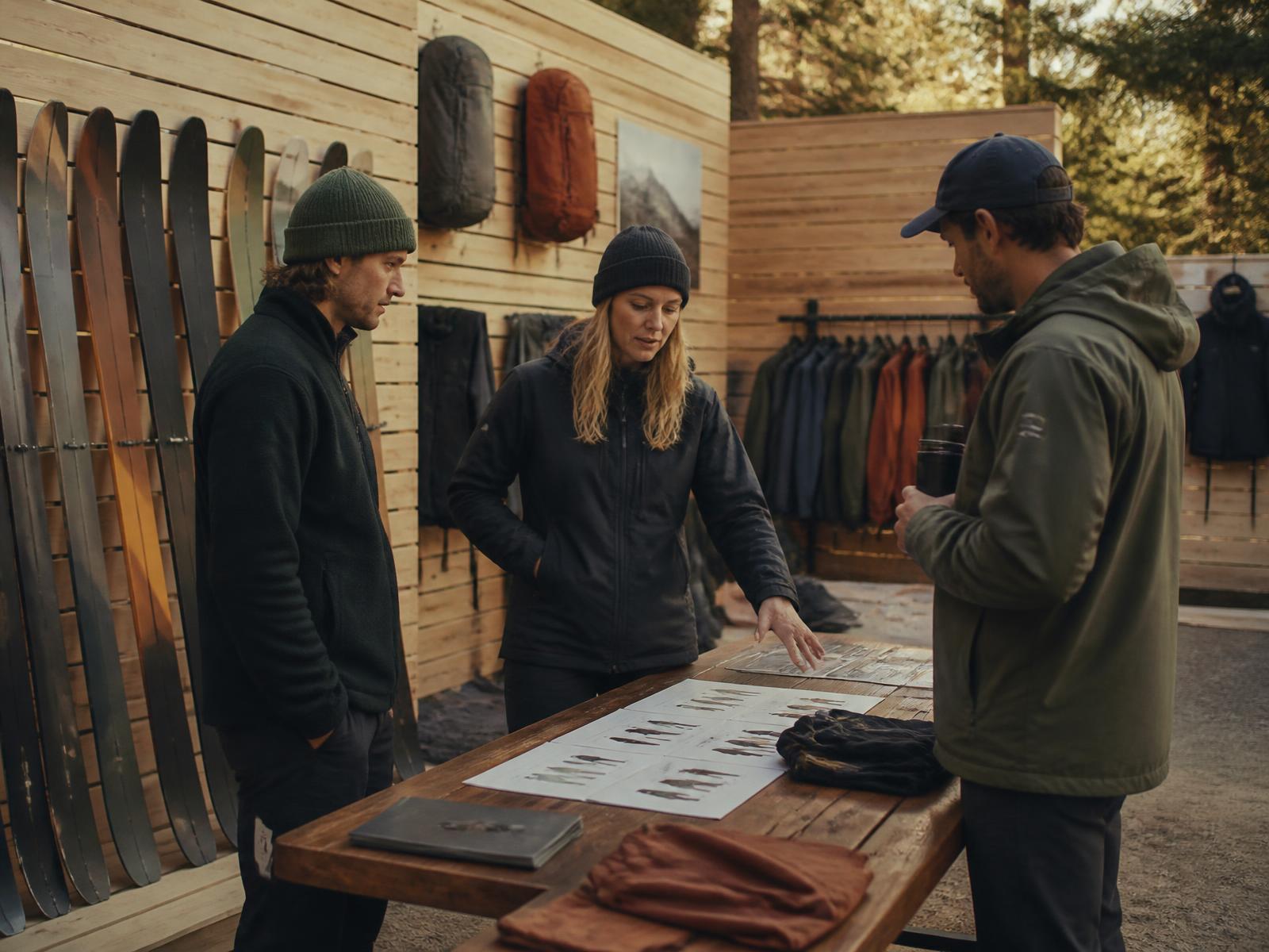Two brand staff walking a specialty retail buyer through next season's line at a wooden table in an outdoor brand showroom, skis and apparel displayed behind