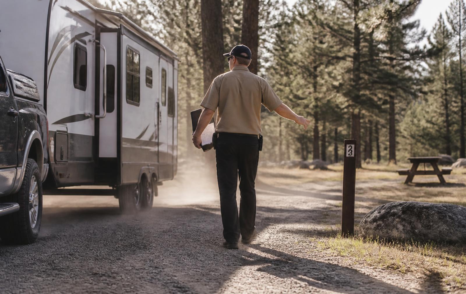 Campground staff member walking beside a travel trailer arriving on a gravel loop, directing it toward a numbered site
