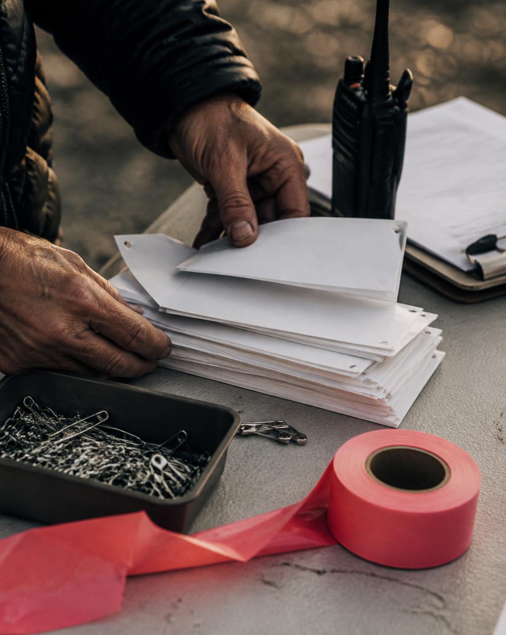 Close-up of hands sorting stacks of blank paper race numbers and safety pins beside a two-way radio, a clipboard and a roll of course-marking tape