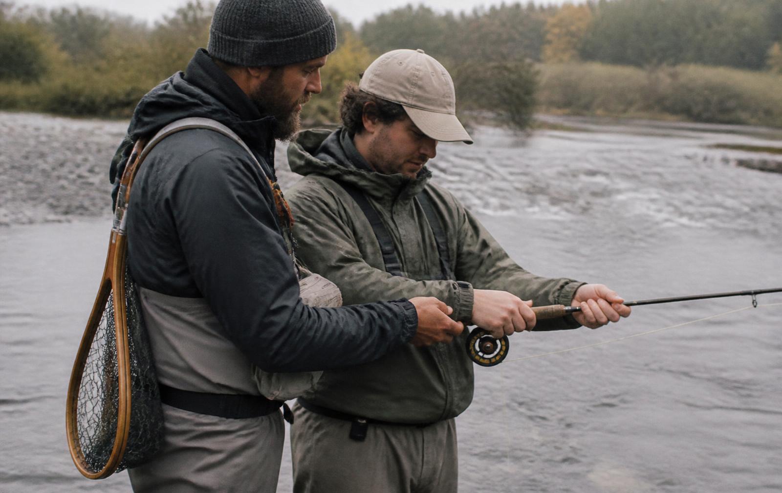 A fishing guide standing beside a client at the edge of a river, showing them how to hold a fly rod, both looking at the rod, a wooden landing net over the guide's shoulder