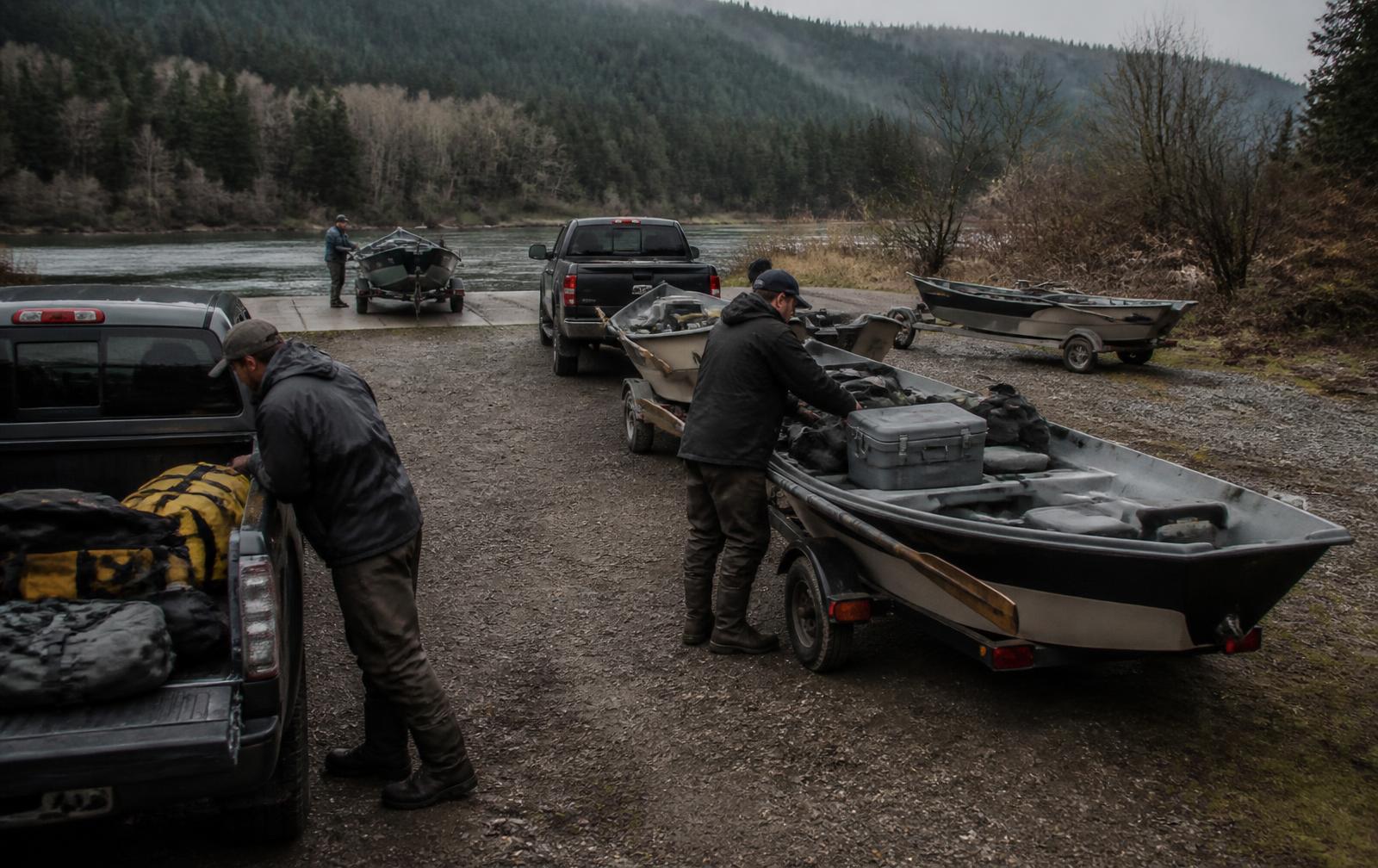 Three drift boats on trailers behind pickup trucks at a river boat ramp in grey morning light, guides loading coolers, dry bags and rod tubes