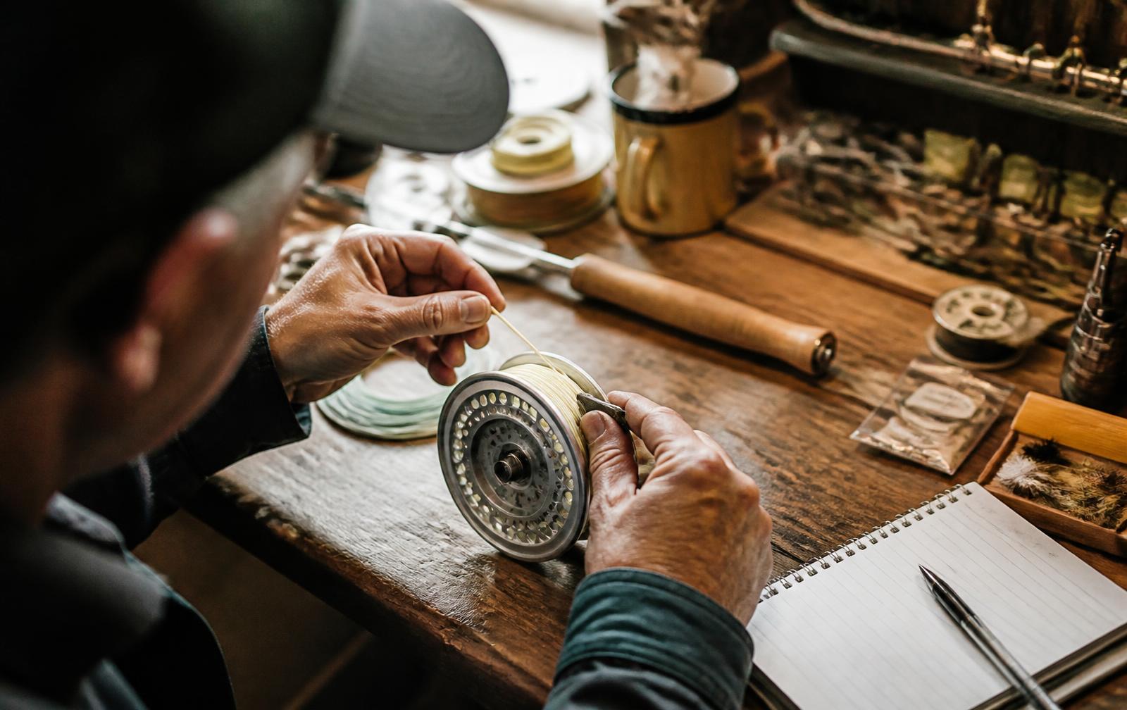 Close overhead view of a fly shop employee's hands winding fly line onto an aluminium reel at a wooden workbench covered with spools, tools and a notebook