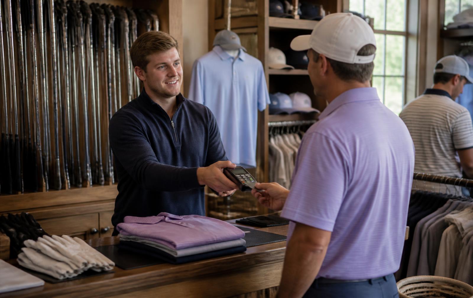 Pro shop staff member handing a card terminal across the counter to a customer buying apparel, with clubs and racks of shirts behind