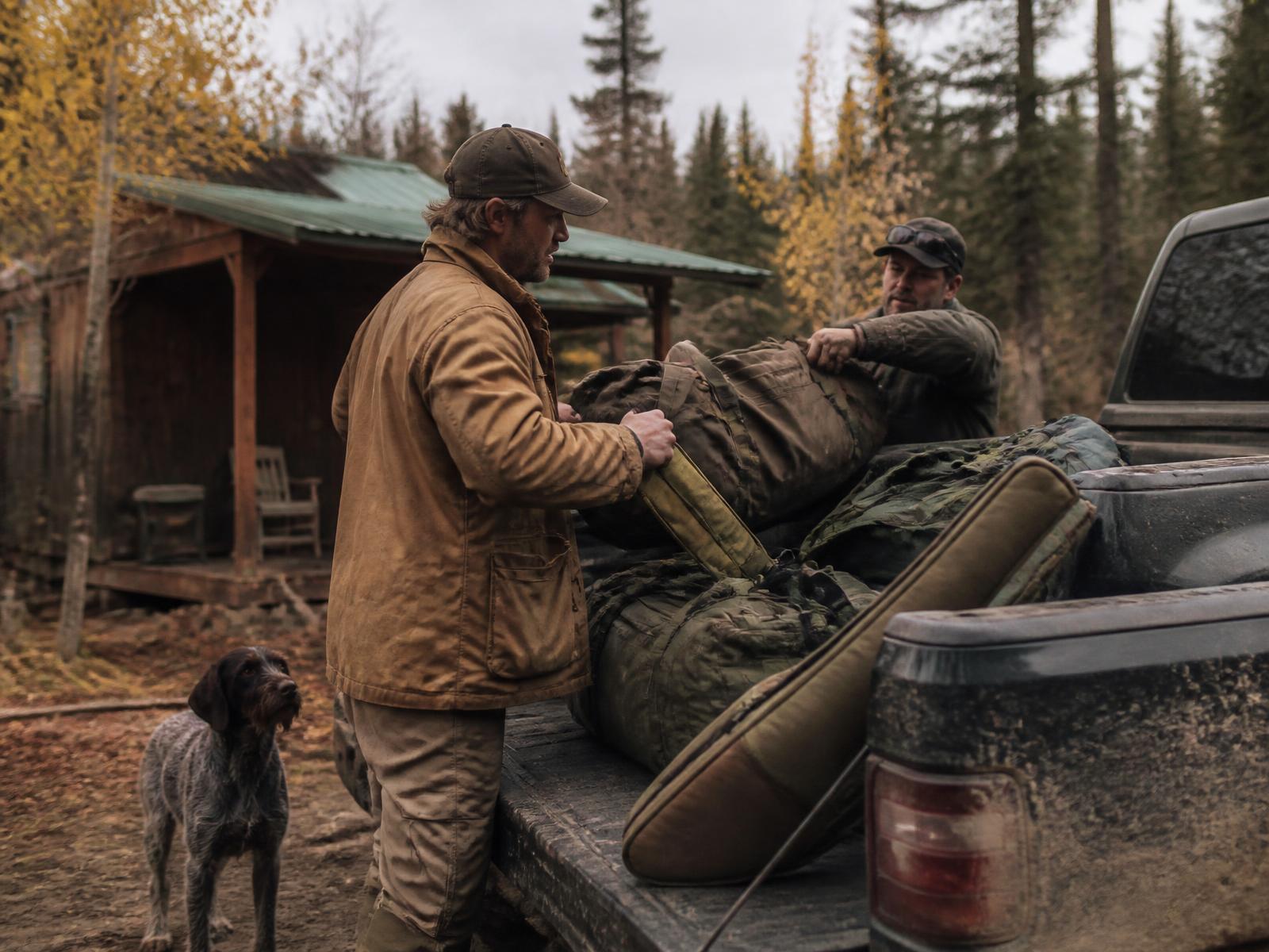 Outfitter and a client loading duffel bags and gear into a pickup outside a backcountry base camp cabin in autumn