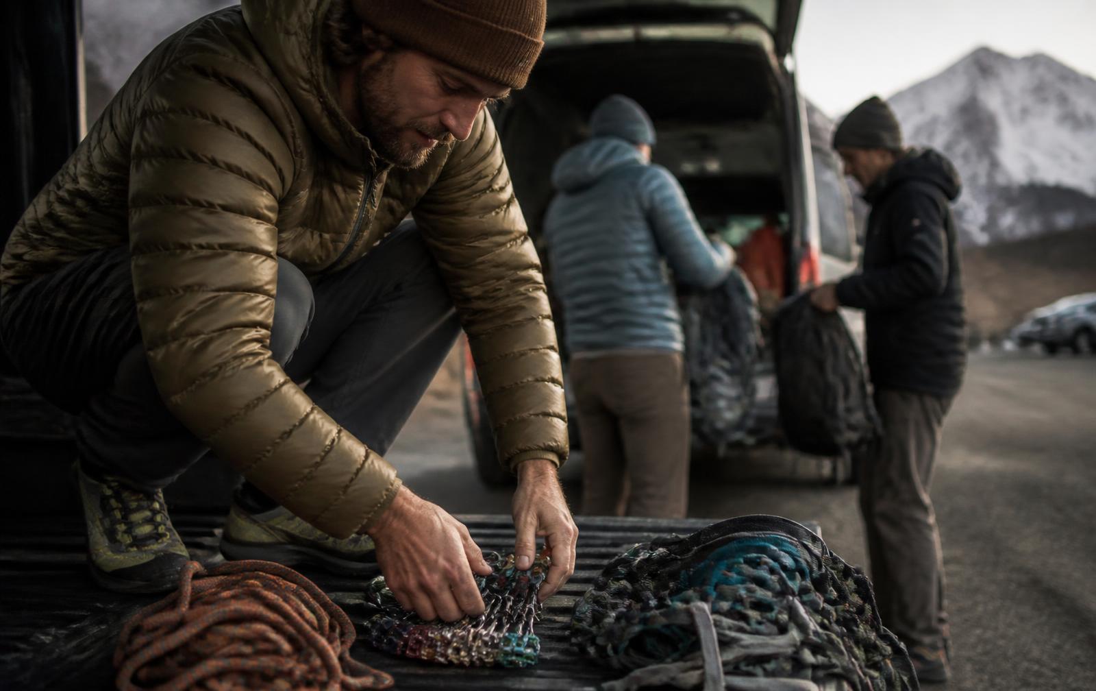 Mountain guide sorting ropes and climbing hardware on a tailgate while clients pull packs from a van at dawn
