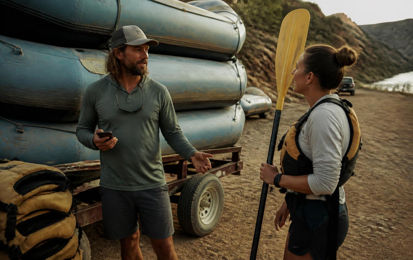 Raft guide holding a phone while talking with a customer beside a trailer stacked with rafts at a riverside pullout