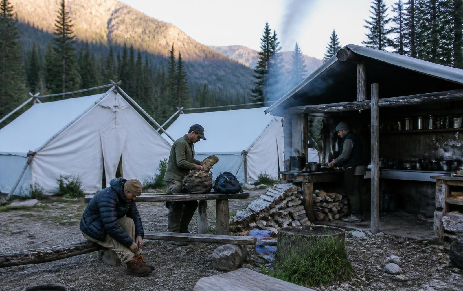 Staff at a remote backcountry camp of white canvas wall tents: one cooking under a timber cook shelter with woodsmoke rising, another organising packs on a bench, a guest lacing boots nearby, forested mountainside behind