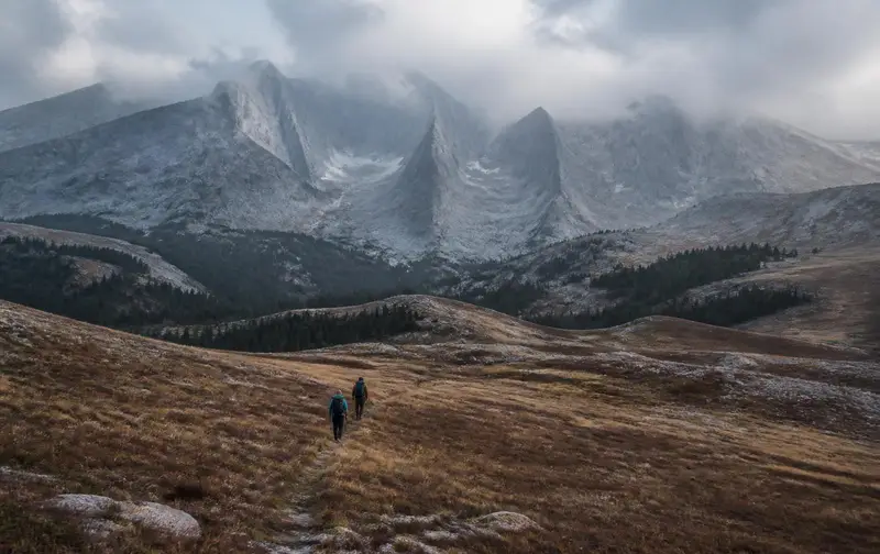 Two small figures with backpacks walking single file along a faint trail across a vast open alpine basin beneath towering grey mountains under low cloud