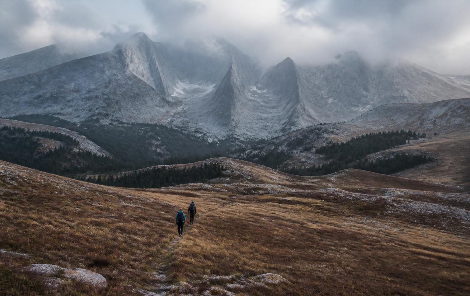 Two small figures with backpacks walking single file along a faint trail across a vast open alpine basin beneath towering grey mountains under low cloud
