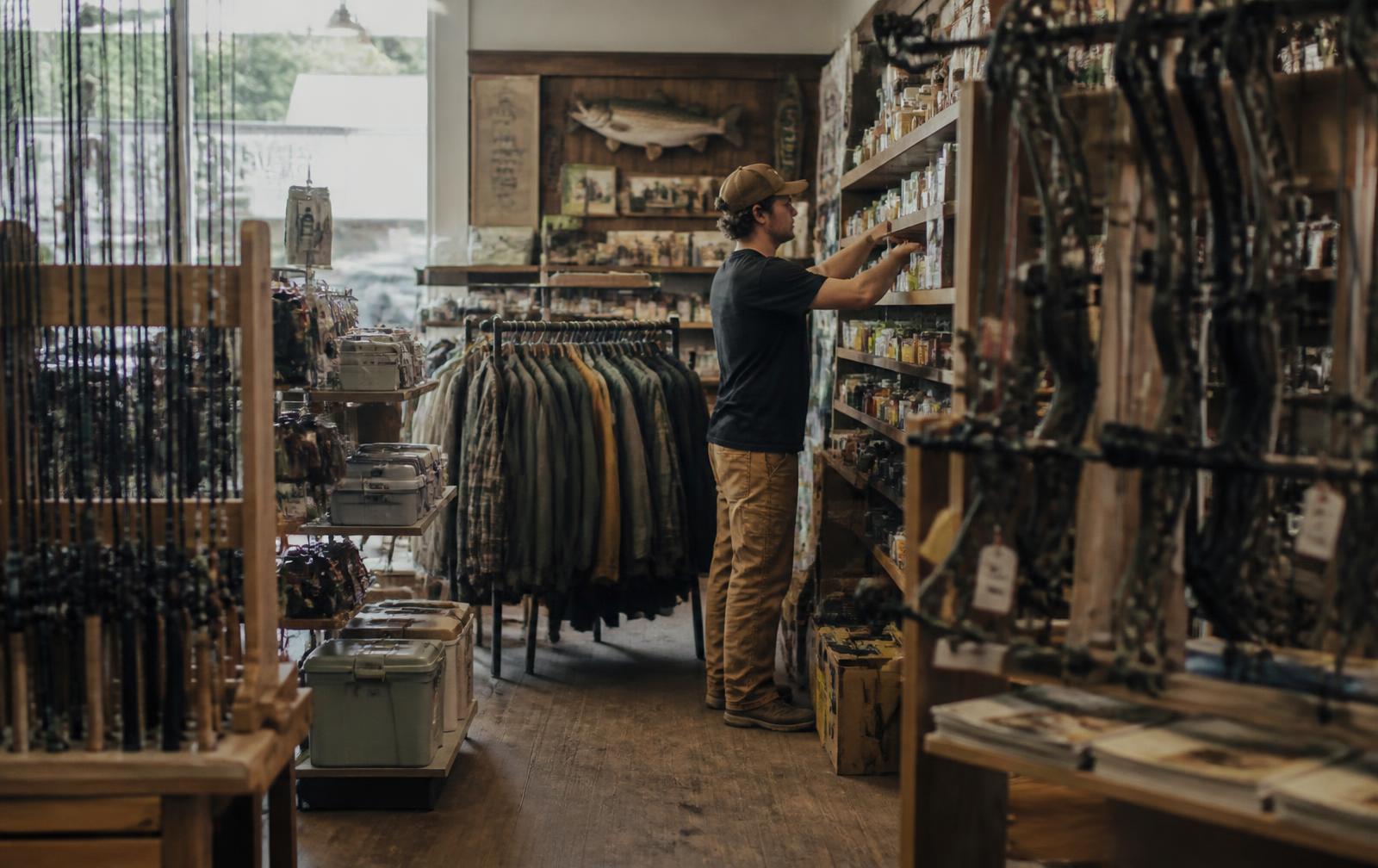 Staff member restocking shelves in an independent outdoor sporting goods store with fishing rods, archery bows and hunting apparel