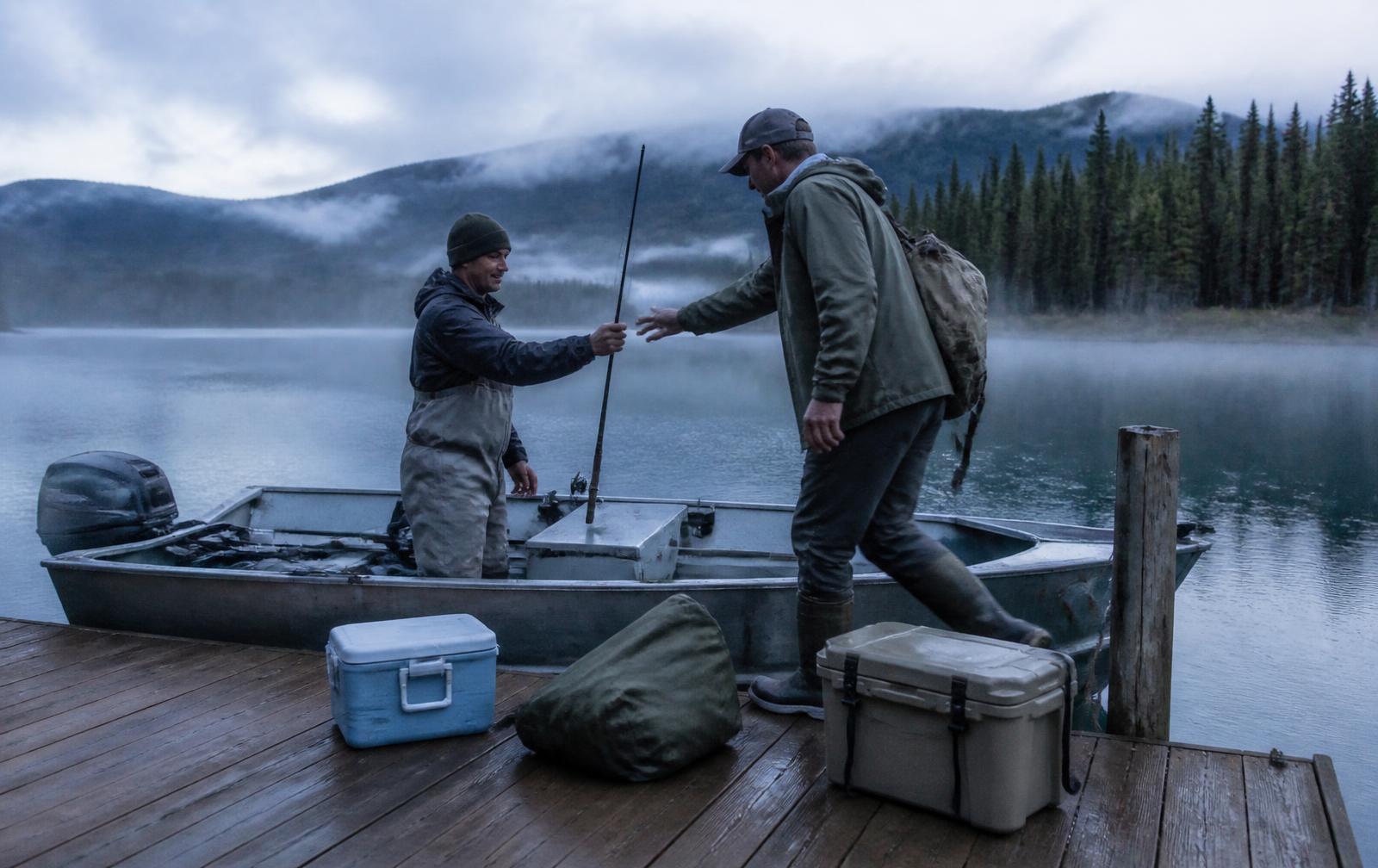 A guide in waders standing in an aluminum boat at a lodge dock at dawn, handing a fishing rod to a guest stepping aboard, mist rising off the lake with forested ridges and low cloud behind, coolers and a dry bag on the wet dock planks