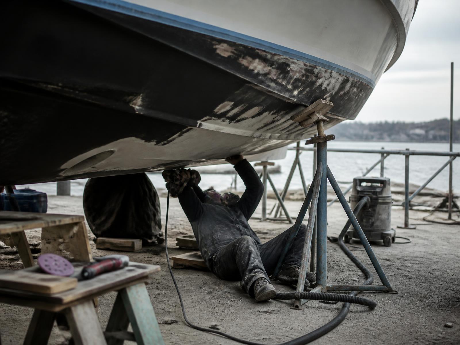 Technician working beneath a boat hauled out on stands in a marine service yard