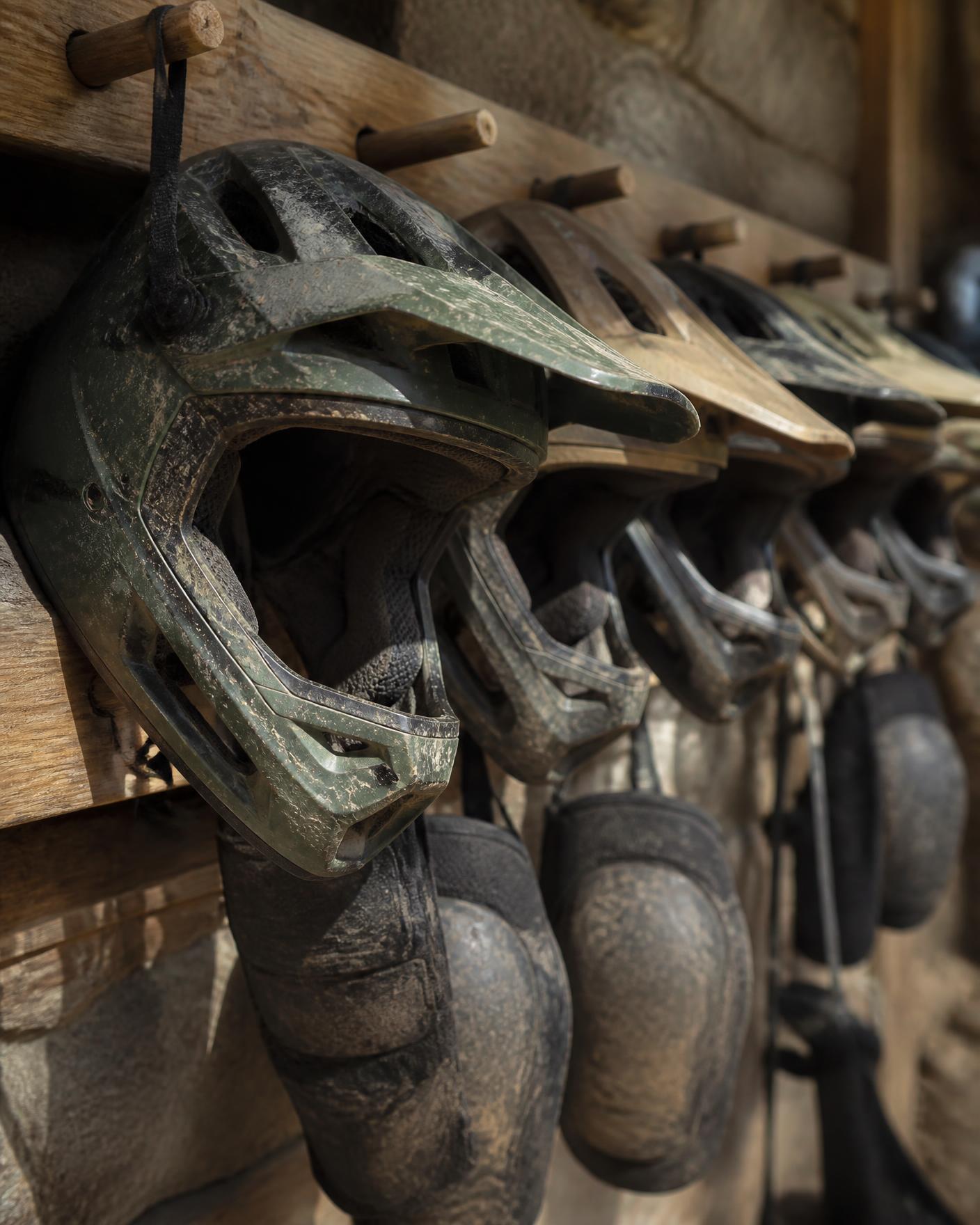 A row of well-used full-face mountain bike helmets and knee pads hanging on worn wooden pegs in a rental shed, raking daylight across scuffed shells and dusty straps