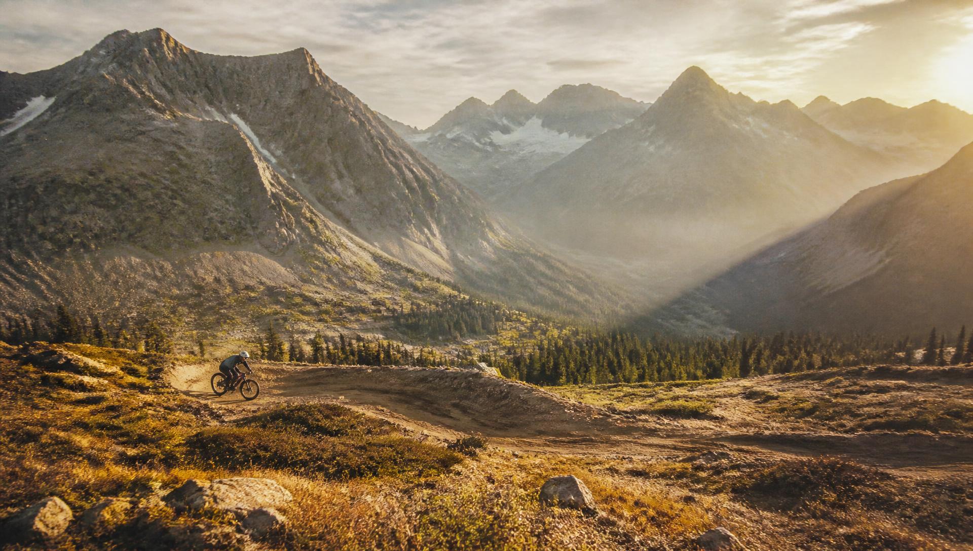 A lone mountain biker small in the frame riding an established dirt trail across an open alpine hillside at sunset, with vast granite peaks, a glacial valley and conifer forest beyond