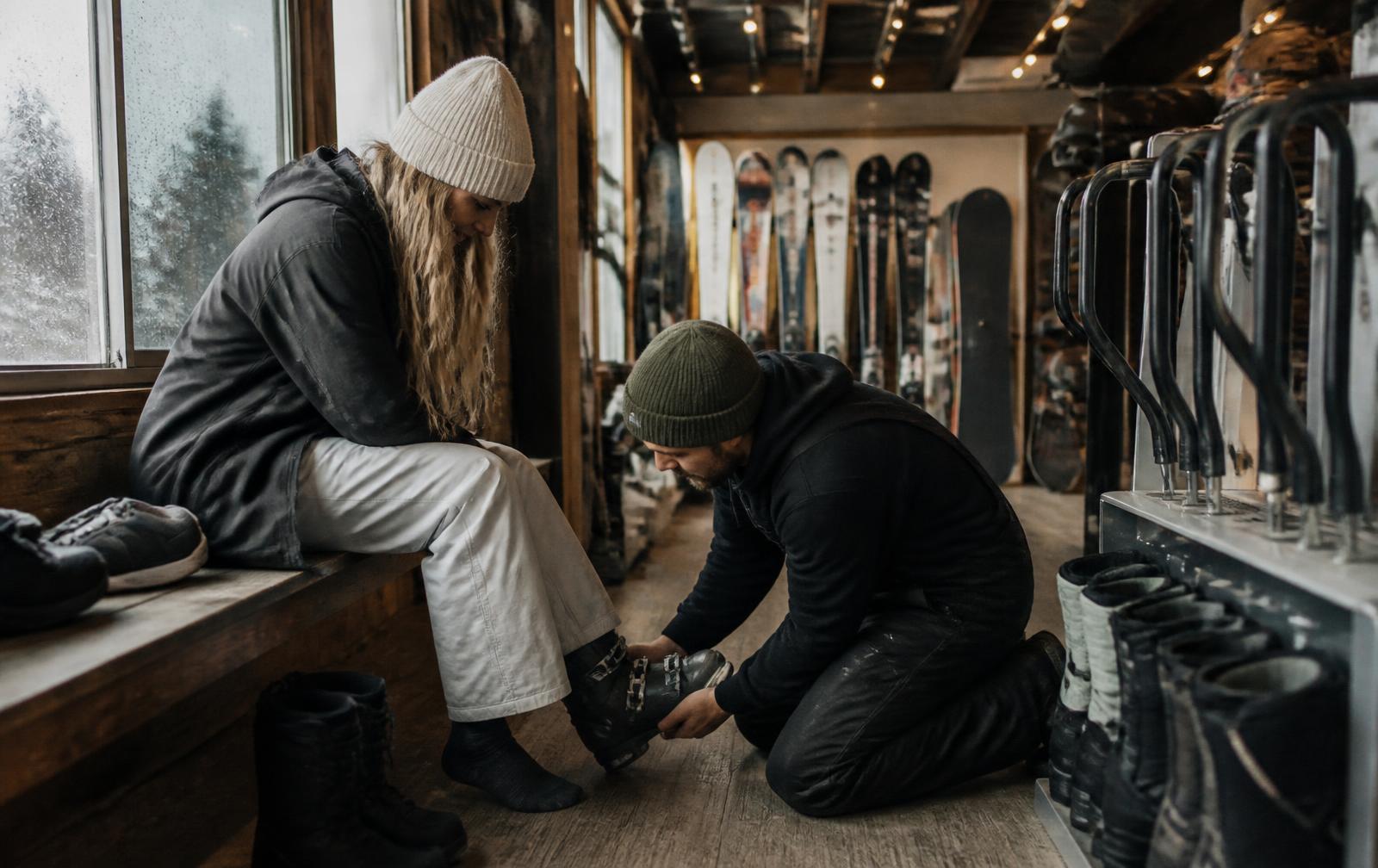 Rental technician kneeling to fit a ski boot for a seated customer in a rental shop lined with skis, snowboards and boot racks