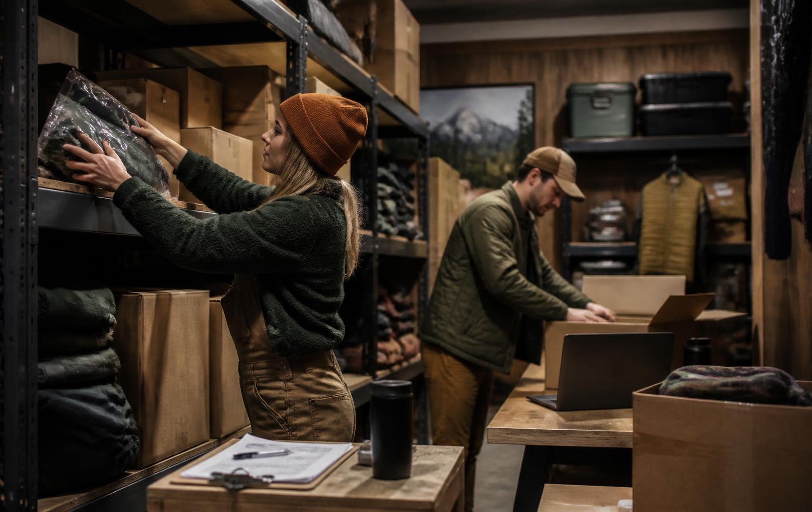 Two staff members in an outdoor retailer's stockroom unpacking new season inventory and packing online orders at a bench