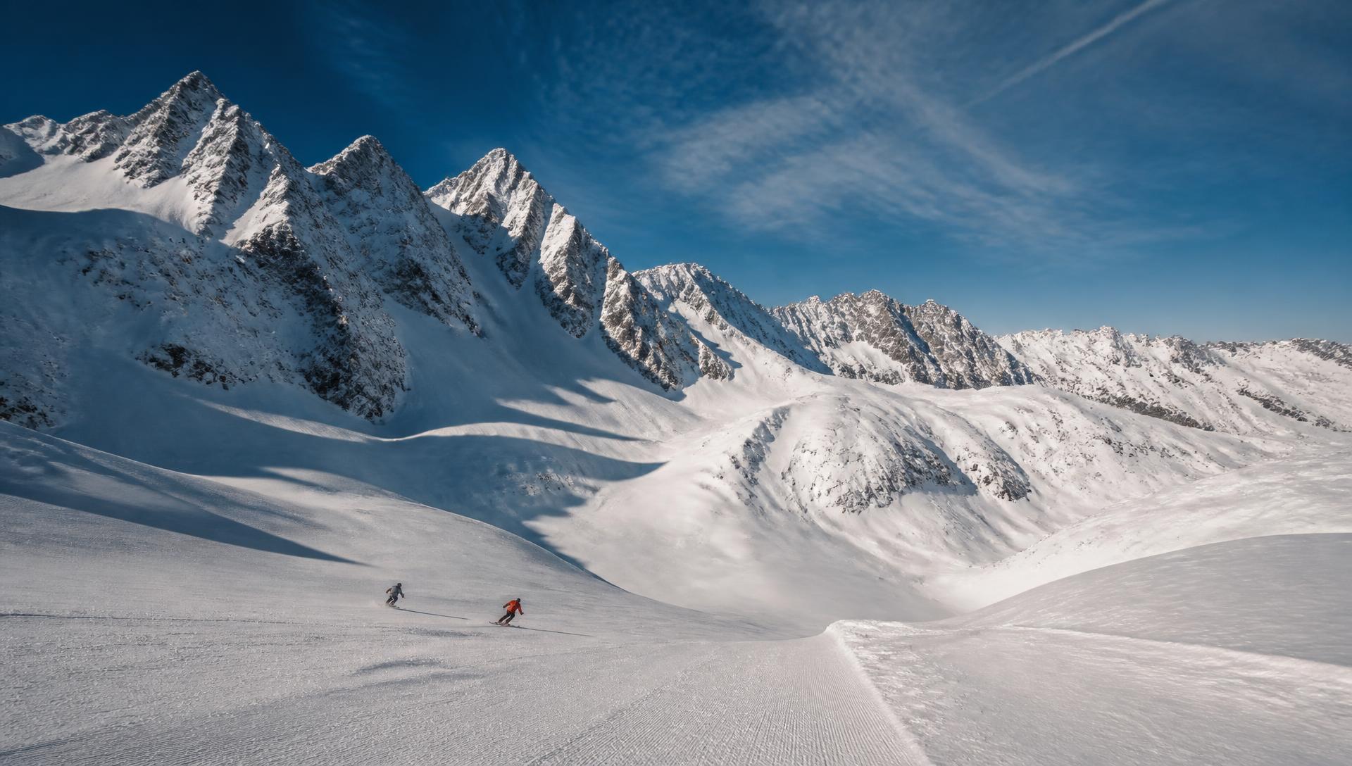 Two small skiers descending a wide groomed run beneath enormous jagged snow-covered peaks and a long alpine ridgeline under a deep blue sky, the landscape dwarfing them