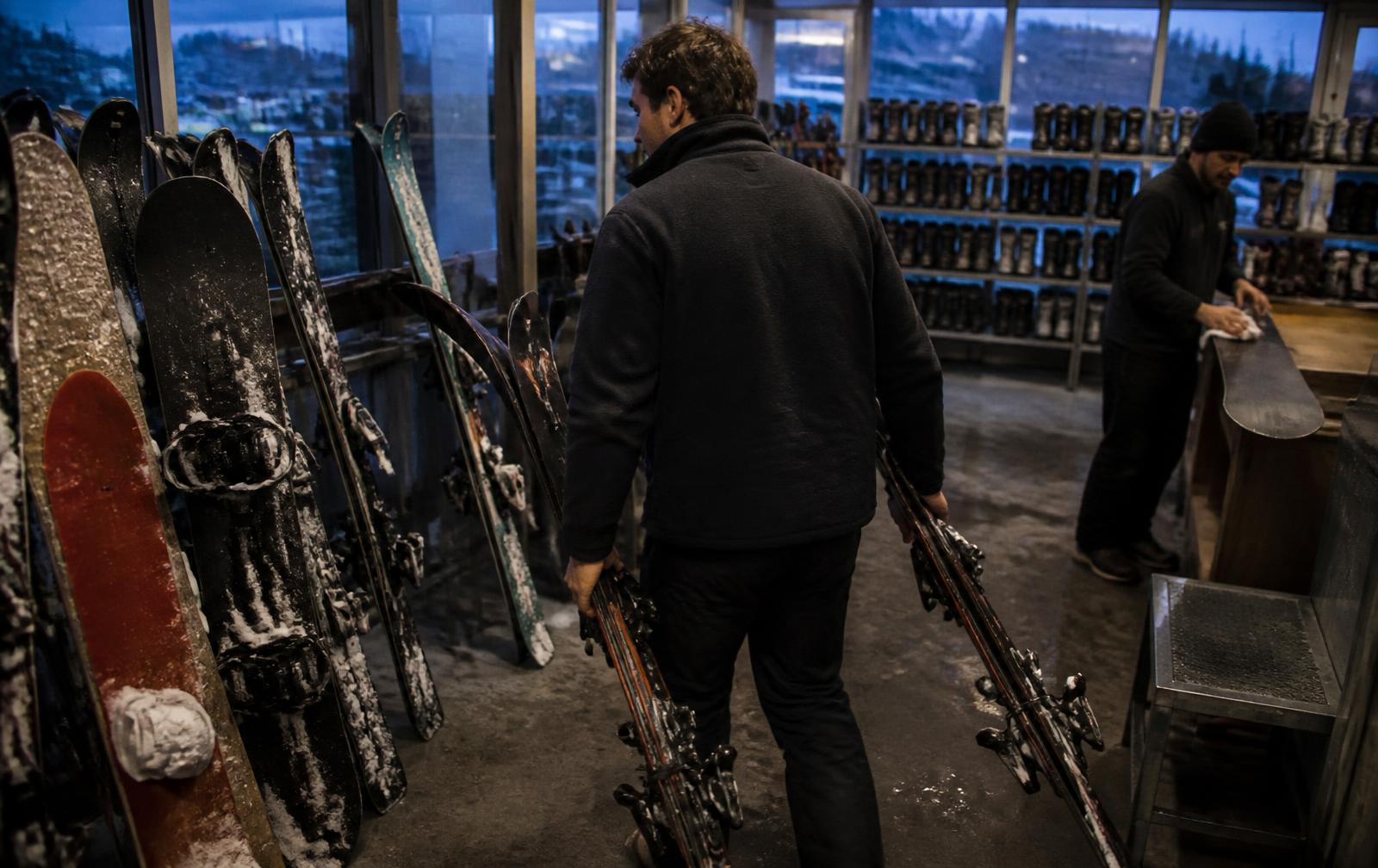 Late afternoon at a ski rental shop return area, a staff member carrying two pairs of snow-covered skis toward the racks while another wipes down a snowboard, rows of returned boards and shelves of ski boots in cold blue window light