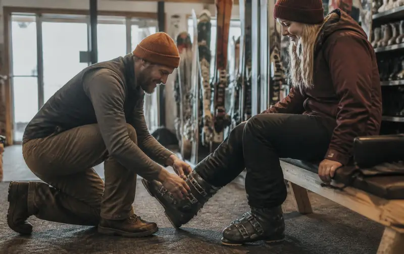 Rental technician kneeling to fit a guest's ski boots inside a resort rental center, with racks of skis and snowboards behind