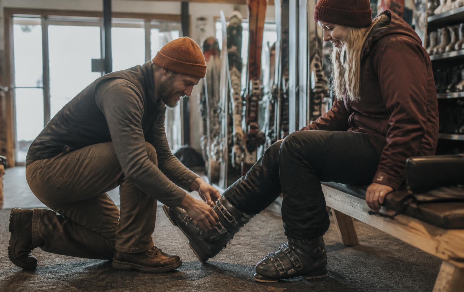 Rental technician kneeling to fit a guest's ski boots inside a resort rental centre, with racks of skis and snowboards behind