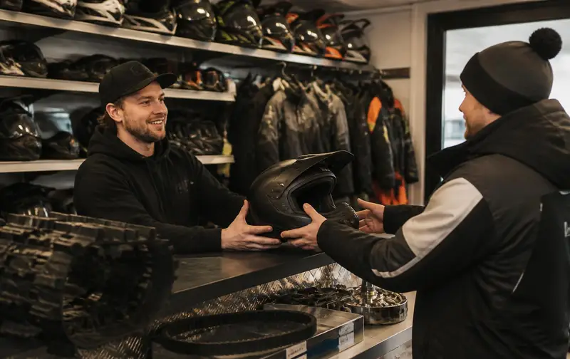 Parts counter employee handing a helmet to a customer, with winter riding suits, goggles and drive belts behind the counter
