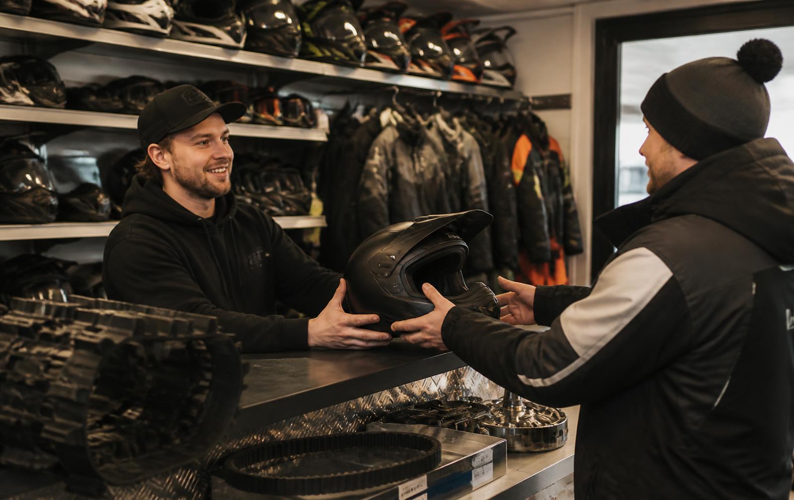 Parts counter employee handing a helmet to a customer, with winter riding suits, goggles and drive belts behind the counter