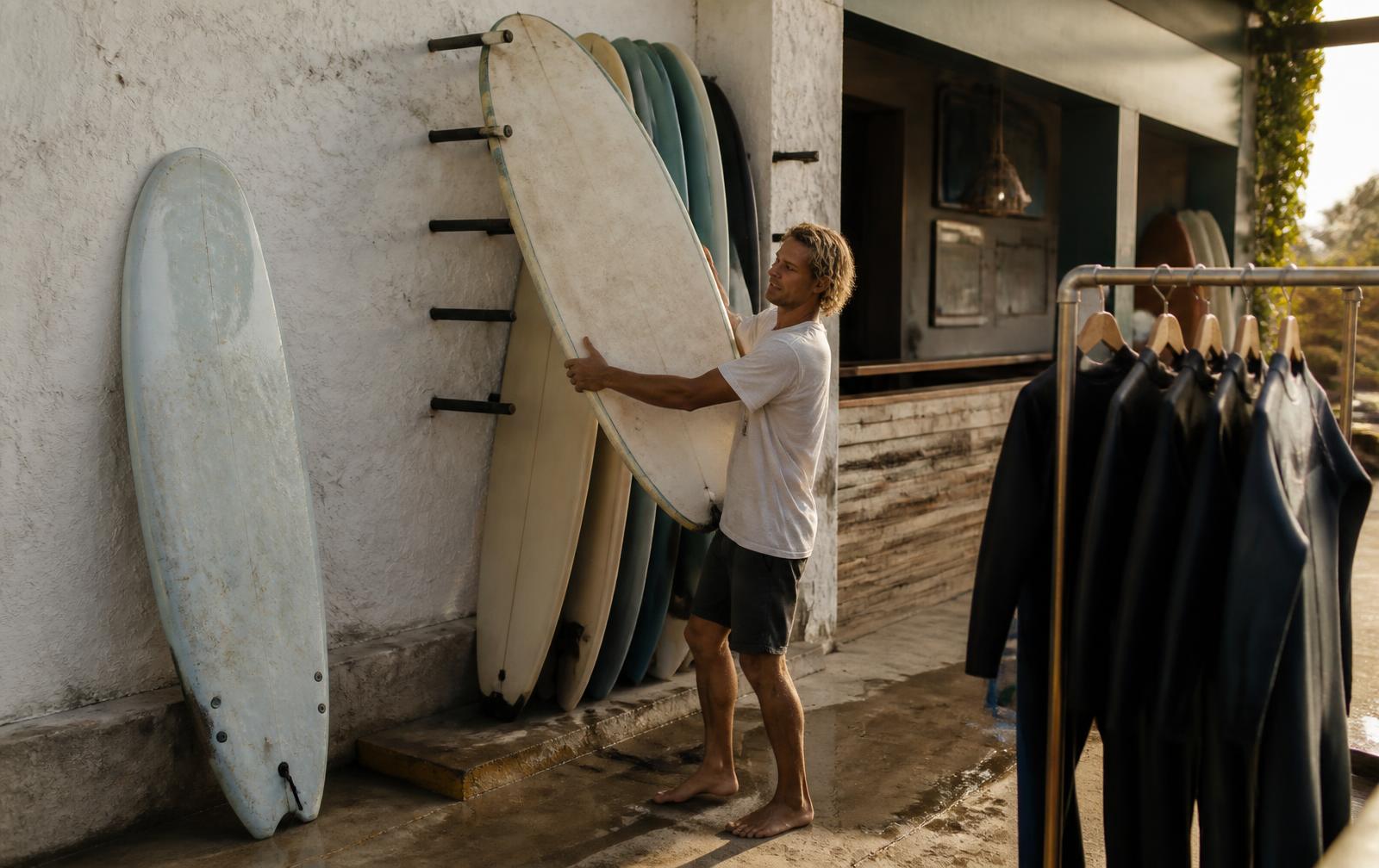 Surf shop employee lifting a soft-top surfboard down from an outdoor rental rack beside a rail of hanging wetsuits in morning light