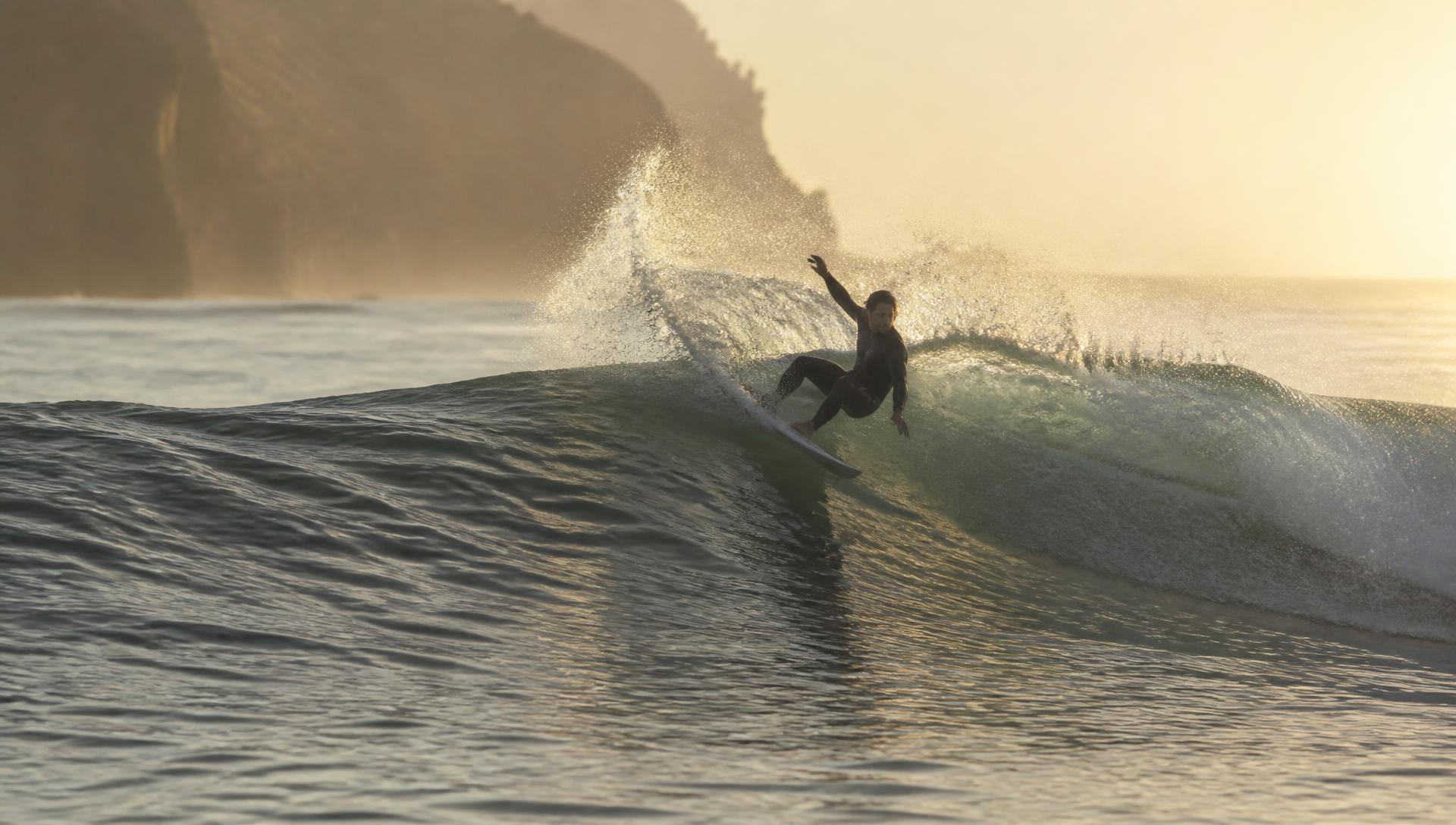Surfer in a black wetsuit carving high on the face of a clean backlit wave at golden hour, spray feathering off the lip with a hazy headland behind